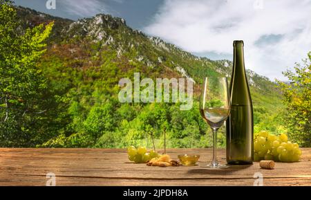 Visita vigneto in estate. Bottiglia e bicchiere di vino bianco in piedi su un tavolo di legno isolato su sfondo montano in serata nuvolosa. Foto Stock