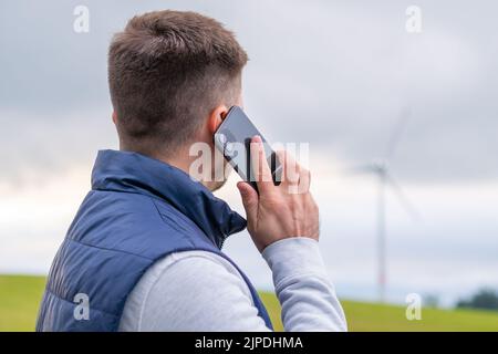 L'ingegnere uomo riferisce circa turbina eolica funzionante via smartphone guardando mulino a vento. La turbina eolica produce energia verde contro il cielo grigio Foto Stock