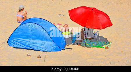 Coppia con il cane dell'animale domestico all'ombra sulla spiaggia sabbiosa di mare che ripartisce il parasole rosso colorful ombrellone & tenda polivalente blu in vacanza soleggiata calda del Regno Unito di luglio Foto Stock