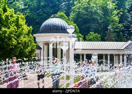 Marienbad, Repubblica Ceca, 30 giugno 2022: La colonata bianca dietro i turisti nascosta dalle gocce sfocate di una fontana Foto Stock