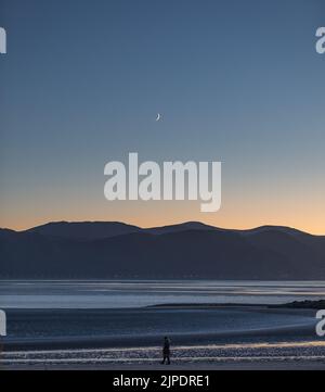 Una coppia non identificabile fa una passeggiata serale lungo la spiaggia vicino al litorale sotto una luna crescente e un cielo blu chiaro. Foto Stock