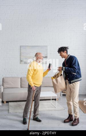 Vista laterale di un pensionato sorridente che tiene uno smartphone vicino al nipote afroamericano con cibo in borsa a casa Foto Stock