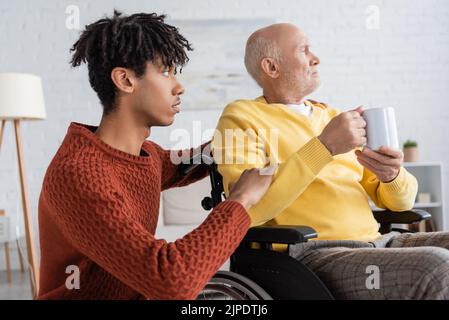Vista laterale del nipote afro-americano che abbraccia il nonno con coppa in sedia a rotelle a casa Foto Stock