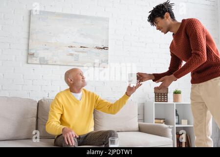 Vista laterale del nipote afro-americano che dà la tazza al nonno sorridente a casa Foto Stock