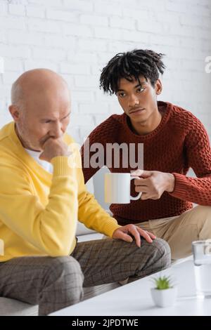 Giovane uomo africano anricano che tiene la tazza vicino nonno dispiaciuto nel paese Foto Stock