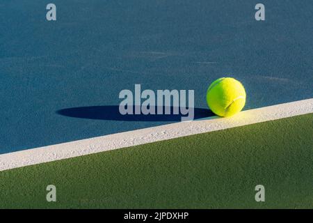 Palla da tennis gialla sulla linea di base su un nuovo campo da tennis blu con verde fuori limite. Foto Stock