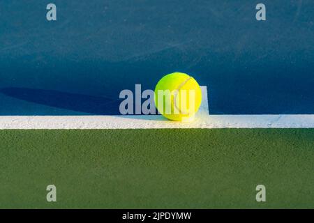 Palla da tennis gialla sulla linea di base al punto centrale su un nuovo campo da tennis blu con verde fuori limite. Foto Stock