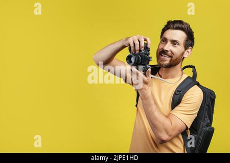 Uomo di mezza età positivo con zaino che fotografa una fotocamera reflex digitale professionale su sfondo giallo, spazio libero Foto Stock