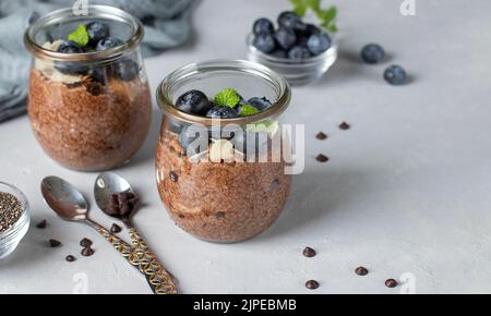 Budino chia al cioccolato con mirtillo e mandorle in vasetti di vetro su fondo grigio chiaro. Cibo sano Foto Stock