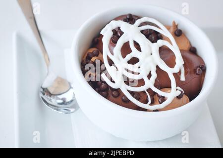 Profiteroles con una guarnizione di lattice di cioccolato bianco in una ciotola di ceramica bianca con cucchiaio d'argento Foto Stock