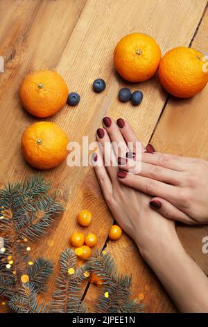Le mani delle donne con manicure di borgogna che giacciono sul tavolo, vista dall'alto. Comfort domestico, tangerini, ramo di abete su sfondo legno. Foto di alta qualità Foto Stock