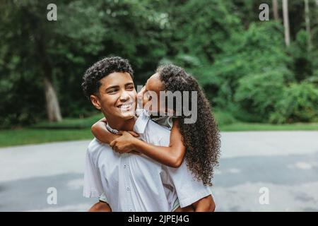 Una sorellina sulla schiena del suo grande fratello adolescente dandogli un bacio sulla guancia in un momento accattivante tra fratelli in famiglia Foto Stock