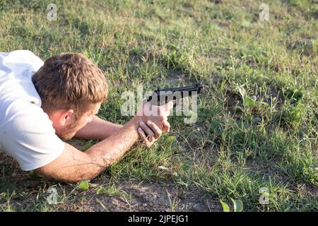 Un uomo caucasico con una pistola pneumatica nelle sue mani punta a un bersaglio, sdraiato sul terreno. Passione per armi e tiro. Spazio copia. Foto Stock