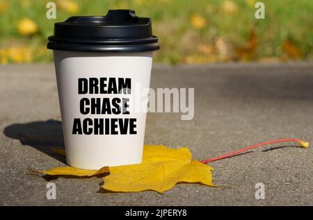 Su una foglia di acero giallo c'è una tazza di caffè su cui è scritto - sogno, inseguimento, realizzare. Concetto aziendale. Foto Stock