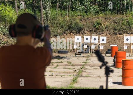 Vista posteriore dell'uomo in cuffie di sicurezza accanto al vetro da campo pratica con pistola a mano sul campo di tiro. Pratica target. Concentrarsi sullo sfondo. Ripresa orizzontale per esterni. Foto di alta qualità Foto Stock