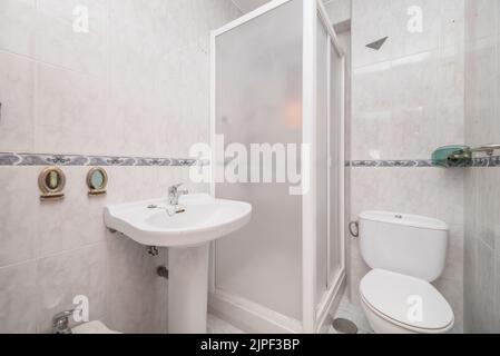 Bagno piastrellato con rifiniture grigie, lavabo in porcellana bianca con piedistallo coordinato e box doccia con rifiniture in alluminio bianco Foto Stock