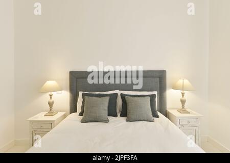 Testiera della camera da letto rivestita in tessuto grigio con cuscini coordinati, due tavoli laterali in legno bianco con lampade e pareti dipinte di bianco sporco Foto Stock