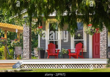 Vista su un grande portico frontale con mobili e piante in vaso. Posti a sedere sul portico rustico in stile rustico con sedie rosse Adirondack e fiori freschi. Foto Stock