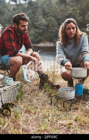 Giovane coppia in campeggio sul lago. Giovane donna che prepara il cibo su una stufa da campeggio con il suo ragazzo seduto vicino. Foto Stock