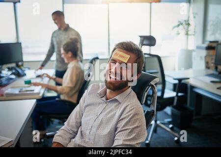 C'è qualcosa sulla mia fronte. Un giovane uomo d'affari allegro con un foglietto appiccicoso sulla sua fronte dicendo essere felice mentre è seduto in ufficio. Foto Stock