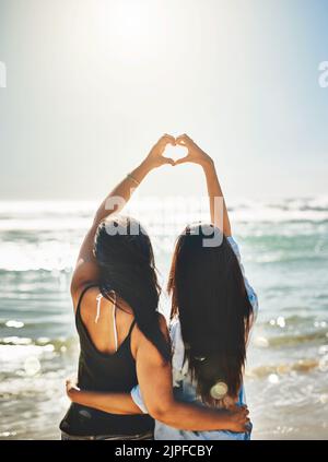 Tutti amano l'estate. Ripresa da dietro di due amici che formano un cuore insieme sulla spiaggia. Foto Stock