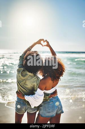 L'estate è qui. Ripresa da dietro di due amici che formano un cuore insieme sulla spiaggia. Foto Stock