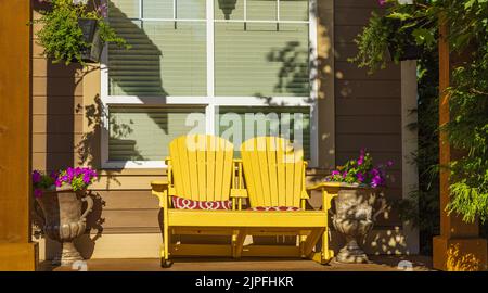 Vista su un grande portico frontale con mobili e piante in vaso. Portico rustico in stile rustico con sedie gialle Adirondack e fiori freschi Foto Stock