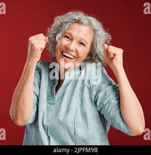 Festeggia, rallegrati e vinci con una donna anziana felice, sorridente ed eccitata che si presenta in studio su uno sfondo rosso. Ritratto di un allegro Foto Stock