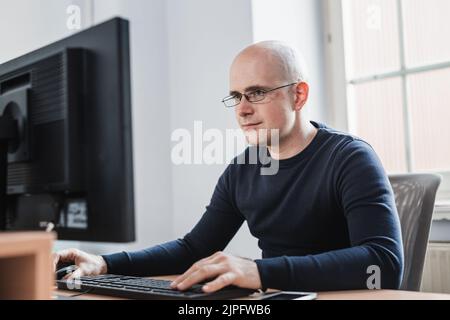 Giovane imprenditore che indossa occhiali seduti al suo tavolo all'interno dell'Ufficio, guardando il rapporto sullo schermo del suo computer. Foto Stock