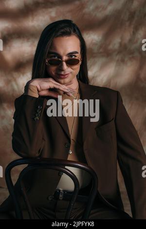 Ritratto di una persona sorridente in occhiali da sole e giacca guardando la macchina fotografica mentre si siede sulla sedia su sfondo marrone astratto Foto Stock