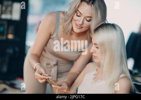 Parrucchiere femmina che fa estensioni di capelli a giovane donna con capelli biondi in salone di bellezza. Prolunga professionale per capelli Foto Stock