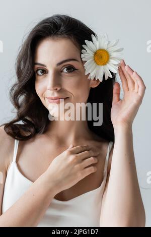 Ritratto di donna sorridente con trucco che tocca camomilla nei capelli e guardando la macchina fotografica isolata sul grigio Foto Stock