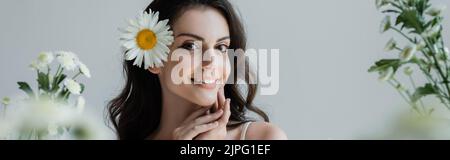 Giovane donna con makeups e camomilla in capelli sorridendo alla macchina fotografica vicino fiori sfocati isolato su grigio, banner Foto Stock