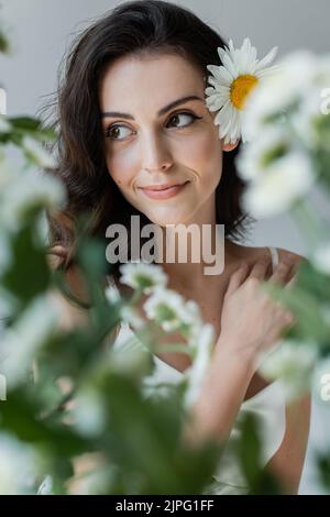 Donna sorridente con visage e camomilla nei capelli che guarda lontano vicino a piante sfocate isolate sul grigio Foto Stock