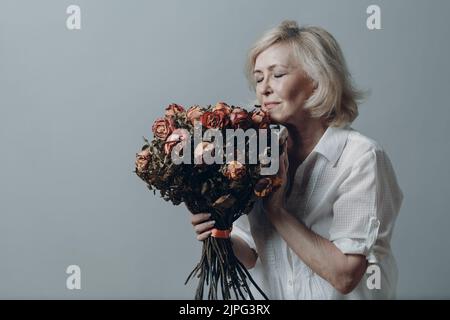 Triste donna anziana che tocca bouquet di fiori di rosa appassiti. Donna matura con capelli grigi in depressione. Concetto di invecchiamento vecchio. Riprese in studio isolate su sfondo grigio. Spazio di copia Foto Stock