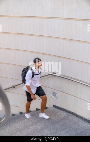 Uomo ispanico vestito in abito casual con cappuccio all'indietro che cammina sulle scale con zaino in città. Foto Stock