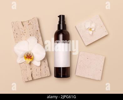 Bottiglia cosmetica in vetro scuro con etichetta vuota vicino a fiori di orchidea bianca e pietre su giallo chiaro, vista dall'alto, mockup. Prodotti di bellezza fatti a mano per la cura della pelle, Foto Stock