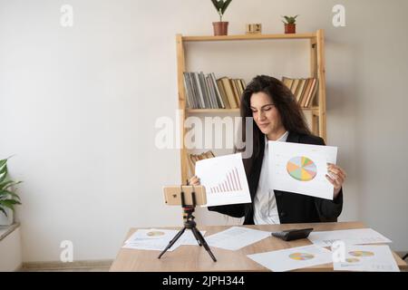 Una donna d'affari positiva che registra contenuti video per il suo blog in ufficio, podcast online in streaming sul telefono cellulare su esposizioni su treppiede e parla di Foto Stock