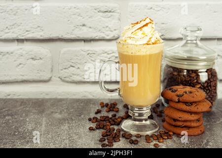 Caffè latte al caramello in un bicchiere alto con panna montata e biscotti al cioccolato. Chicchi di caffè sulla superficie della parete in mattoni bianchi e grigi con spazio per le copie. Foto Stock