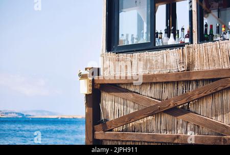 Foto esterna di un cocktail bar sulla spiaggia di giorno che mostra una gamma di bottiglie di alcool attraverso la finestra aperta Foto Stock