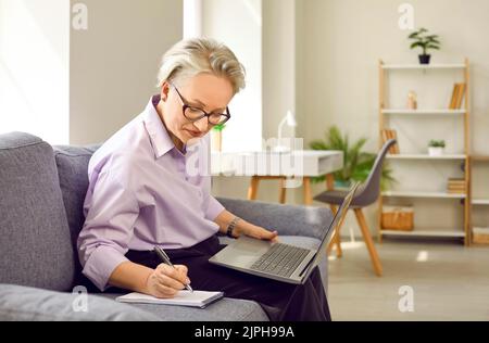 Donna anziana dai capelli grigi con occhiali siede sul divano con il laptop in grembo e prende appunti nel notebook Foto Stock