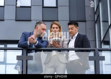 Tre colleghi, diversi gruppi aziendali, persone aziendali al di fuori dell'edificio dell'ufficio che parlano e discutono del processo di lavoro, guardando con attenzione il tablet Foto Stock