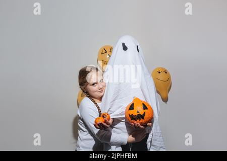 Halloween felice. Una bambina abbraccia un fantasma in un vestito bianco e tiene una zucca arancione. Palline arancioni dipinte. Emozioni diverse di gioia, rabbia, risate. Design festivo, concetto di festa. Messa a fuoco selettiva morbida Foto Stock