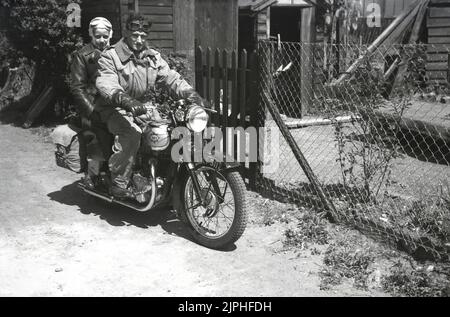 1950s, storico, una coppia seduta su una motocicletta BSA dell'epoca parcheggiata su un sentiero sterrato fuori da un cortile, Inghilterra, Regno Unito. Cappotti pesanti e occhiali da vista, guanti in pelle e panniere in tela sul retro della moto, stanno per andare in giro. Foto Stock