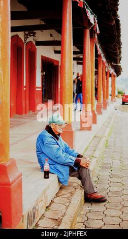 buon vecchio uomo, seduto in attesa, seduto sui gradini mentre si beve una cola, si possono vedere le colonne di una casa rossa Foto Stock