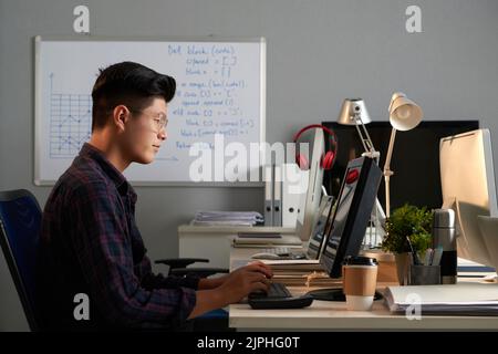 Programmatore asiatico che lavora duro indossando camicia a segno e occhiali seduti alla scrivania dell'ufficio e finendo progetto ambizioso, vista del profilo Foto Stock