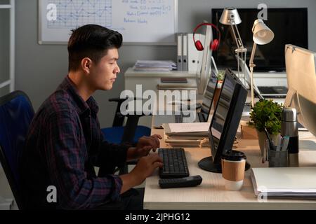 Vista del profilo di un manager asiatico sicuro indossando una maglietta a quadri seduta di fronte al computer moderno e lavorando al progetto di avvio, ritratto Foto Stock