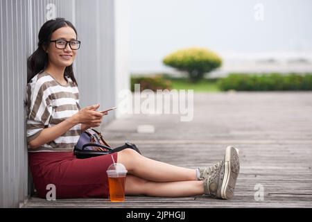 Ritratto completo dell'attraente designer asiatico che si propone per la fotografia con un sorriso tondo mentre si prende una breve pausa dal lavoro e si gode una calda giornata all'aperto Foto Stock