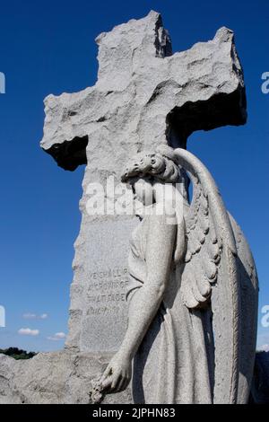 Un angelo di pietra si alza con la mano sulla croce e le sue ali si spalancano parzialmente con uno sguardo di tristezza e perdita contro il cielo blu scuro Foto Stock