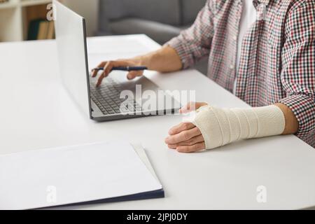 Uomo con fasciatura avvolto intorno al polso ferito seduto alla scrivania e utilizzando un computer portatile Foto Stock